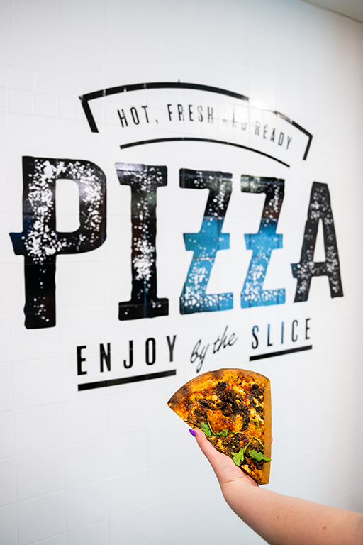 A photo of a wall wrap that says "Hot, Fresh and Ready. Pizza, Enjoy By the Slice" with a slice of pizza held by a person in front.